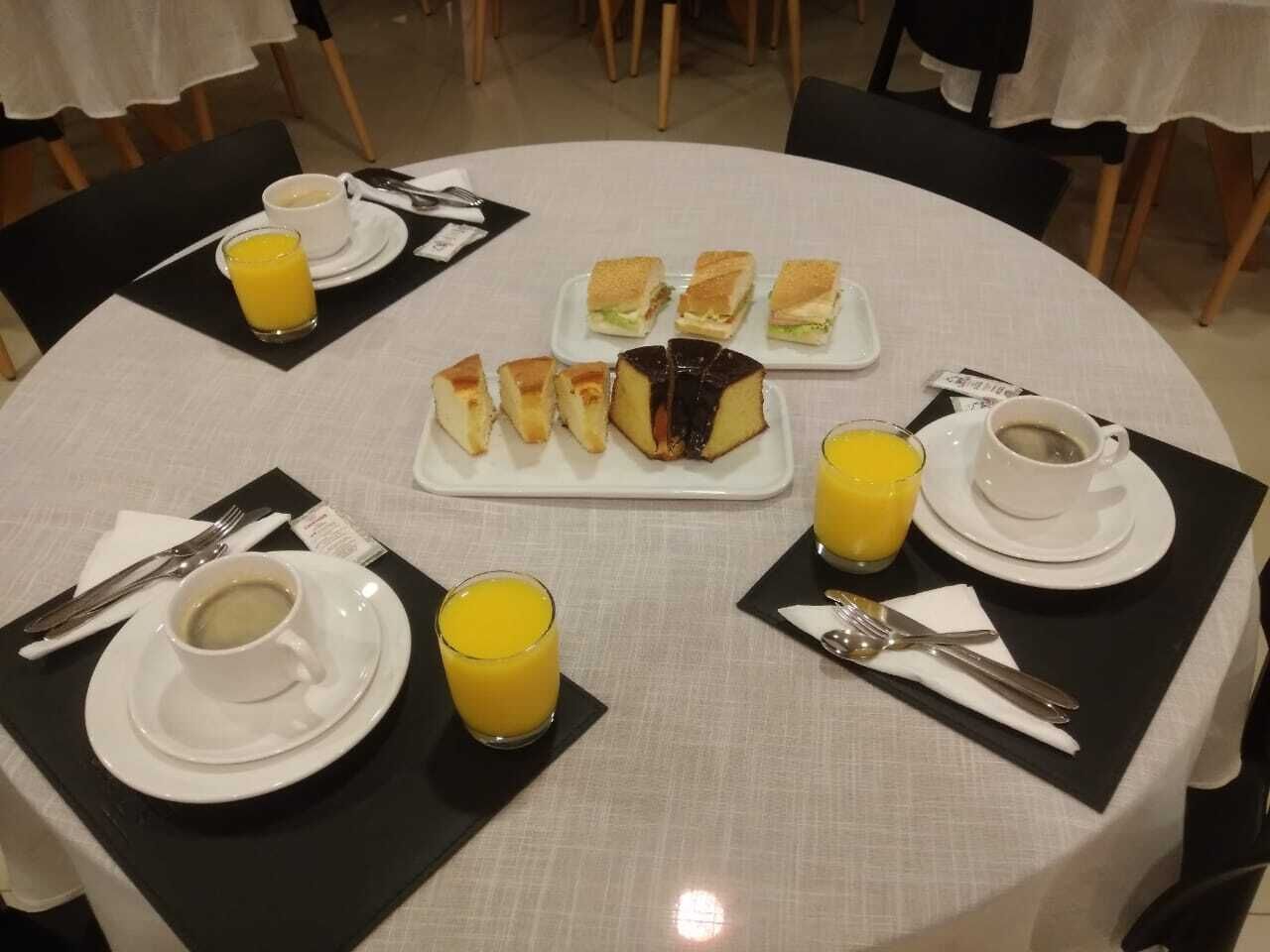 Uma mesa de jantar redonda com toalha branca, posta com pratos, xícaras e talheres. Há vários sanduíches quadrados e uma fatia de bolo sobre a mesa.