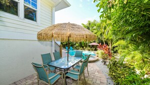 A patio with a table and chairs, a thatched umbrella, and a pool area surrounded by lush greenery. - Beach Time AMI by AMI Locals (Holmes Beach)