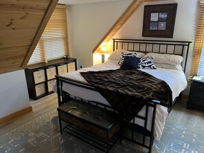 Un dormitorio con cama, mesitas de noche, un banco, un cuadro en la pared y techo inclinado.