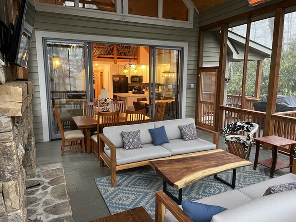 Un espacio exterior acogedor con sofá, mesa de madera y sillas, comunicado a una cocina y comedor mediante puertas de vidrio.