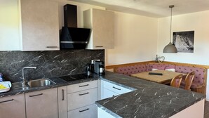 Une cuisine moderne avec un poêle noir, un évier et un plan de travail en marbre. On aperçoit, à l’arrière-plan, un banc en bois recouvert d’une nappe à carreaux.