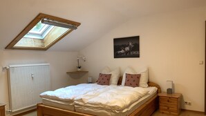 Une chambre avec un plafond en pente, un puits de lumière, un radiateur, une table de chevet, une commode, un cadre de lit en bois et un tableau accroché au mur.