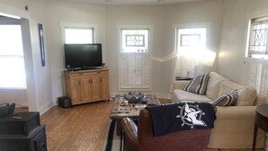 Un salon avec un plancher en bois, un canapé en cuir marron, un divan beige, une table basse, un téléviseur posé sur un meuble en bois et des fenêtres équipées de volets.