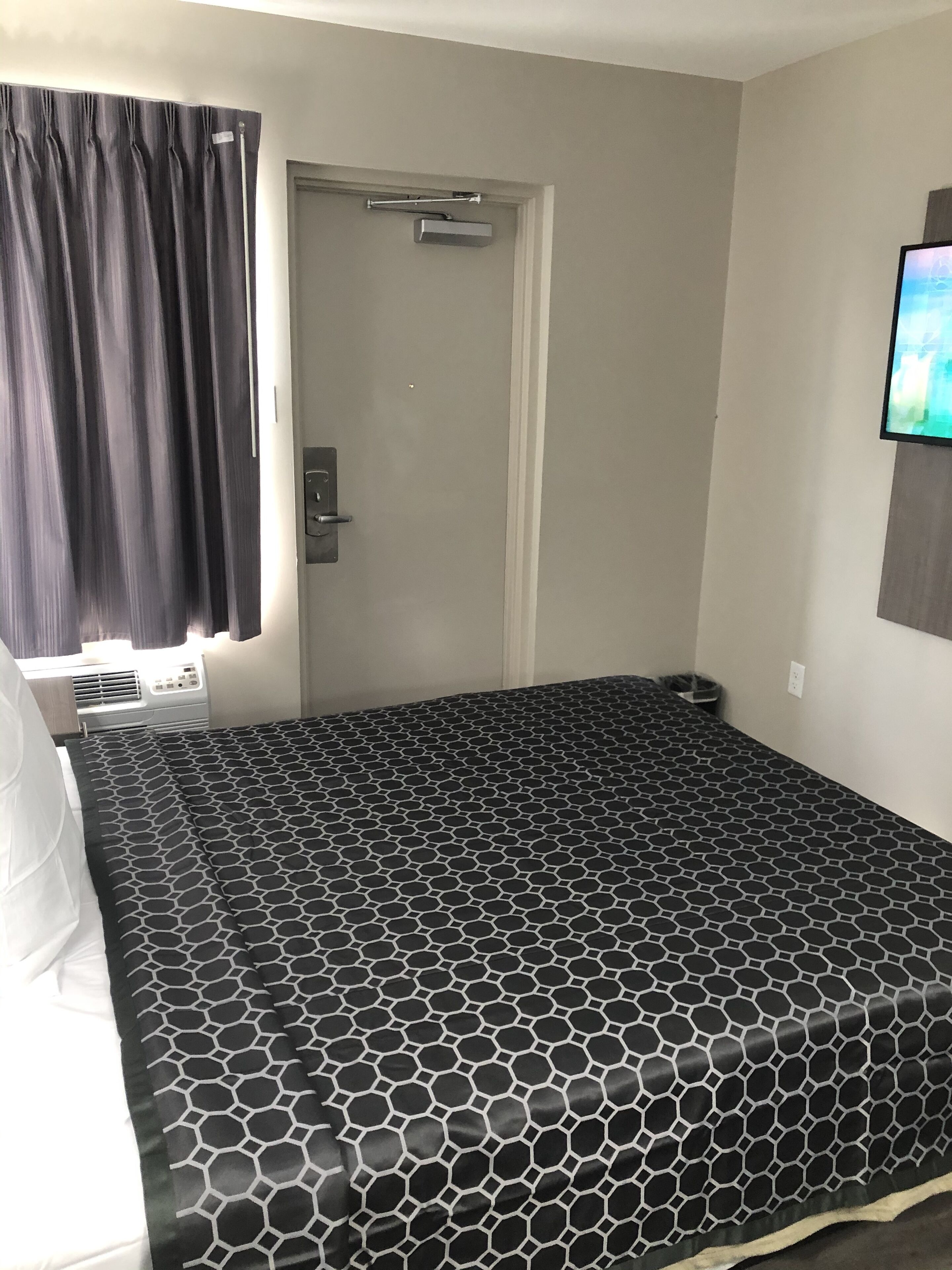 Quarto com cama, porta, janela com cortinas e televisão presa na parede.
