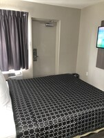 Quarto com cama, porta, janela com cortinas e televisão presa na parede.