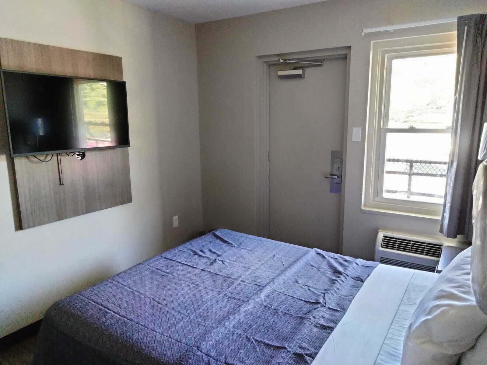 Una habitación de hotel con una cama, un televisor de pantalla plana montado en la pared, una puerta y una ventana con cortinas.