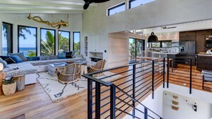 Amplia sala de estar con techos altos, ventanas grandes, cocina moderna y un balcón con vista al océano.