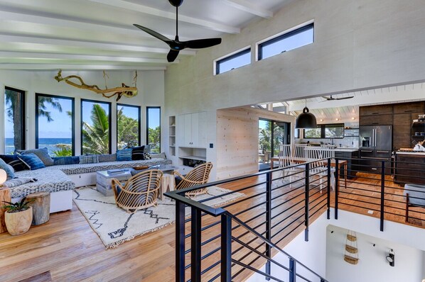 A spacious living area with a high ceiling, large windows, a modern kitchen, and a balcony overlooking the ocean. - Beachfront, Modern Home, Just Steps to the Sand (Haena)