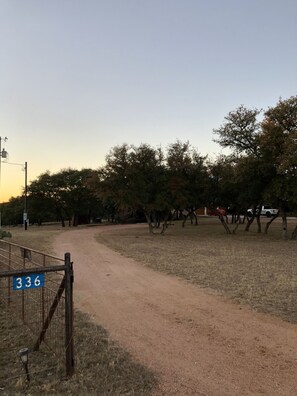 Uma trilha de terra serpenteia por uma área seca e gramada, com árvores e um portão de metal com a inscrição '336'.