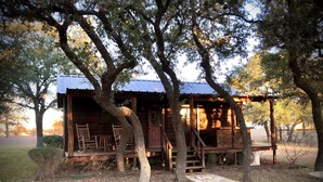A rustic cabin with a porch and wooden furniture, surrounded by trees and a stone pathway. - Cabin in the Country (Fredericksburg)