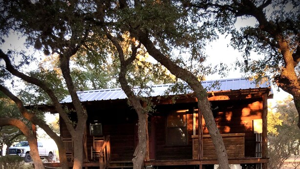 A hot tub with a wooden deck and outdoor seating area, surrounded by trees and a rustic cabin. - Cabin in the Country (Fredericksburg)