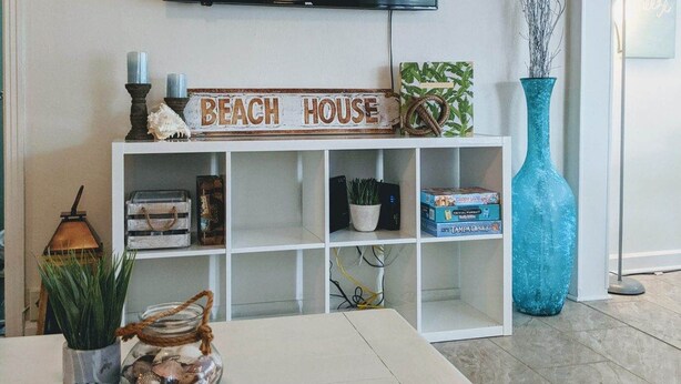A living room with a white console table, a flat-screen TV mounted on the wall, and a sign that reads "BEACH HOUSE". - Tropical Tiny House (New Port Richey)