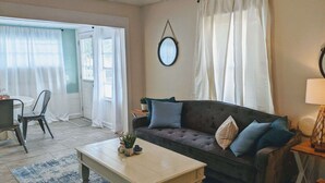 A living room with a sofa, a coffee table, and a chair. There is a window with curtains, a wall-mounted mirror, and a lamp on a side table. - Tropical Tiny House (New Port Richey)