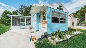 A small, single-story building with a light blue exterior, a covered porch, and a garage. - Tropical Tiny House (New Port Richey)