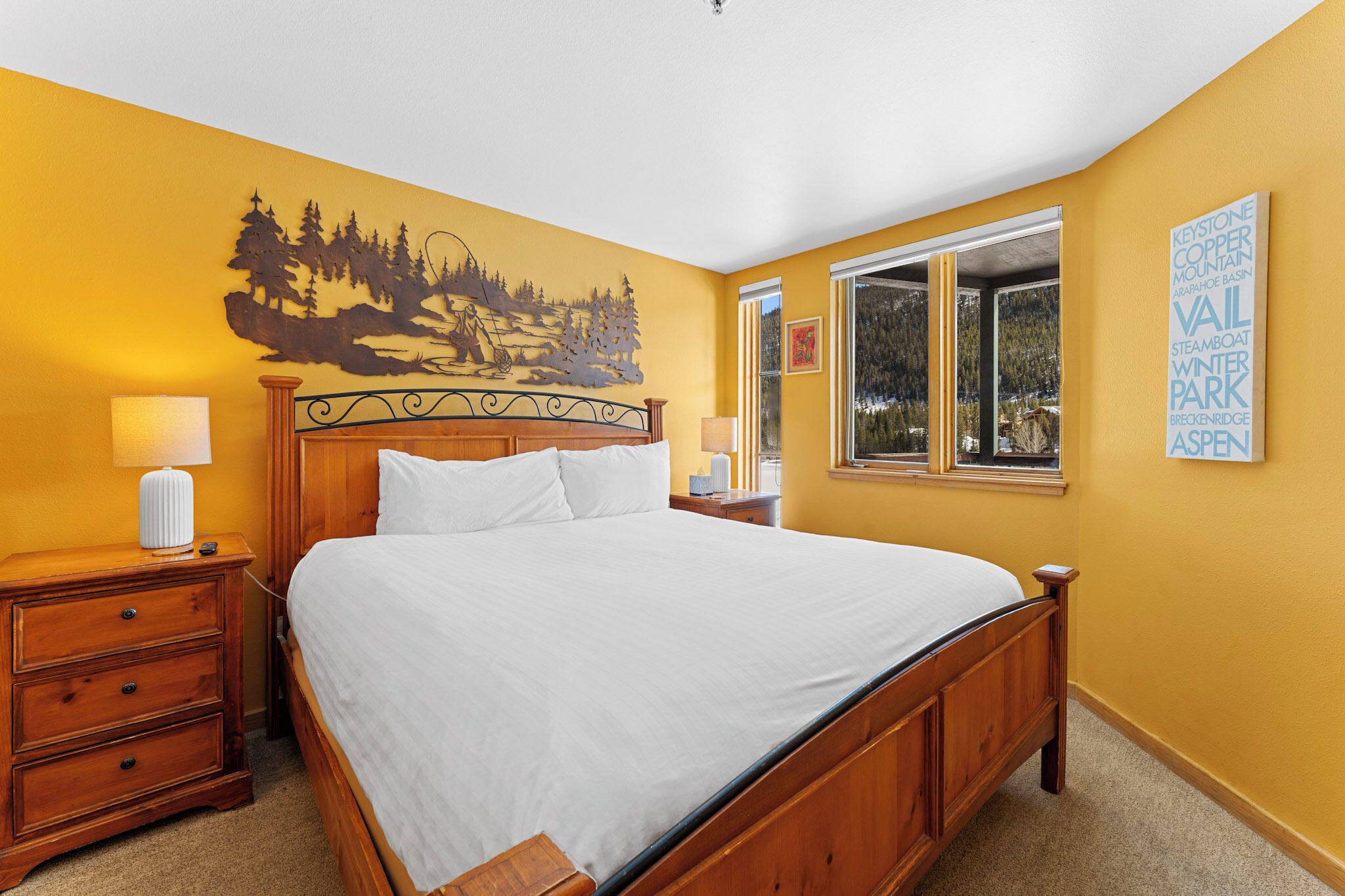 A neatly made bed in a room with a yellow wall, a window with a view, and a decorative metal wall piece.