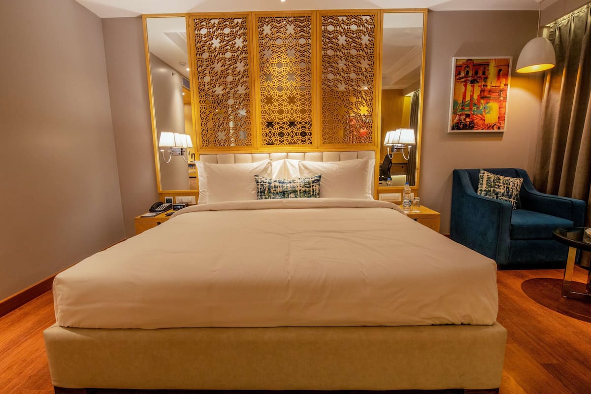 A hotel room with a large bed, a blue armchair, a decorative partition, and a painting on the wall.