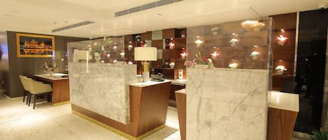 A hotel reception area with marble counters, a reception desk, a seating area with chairs, and a decorative wall with lights and flowers.