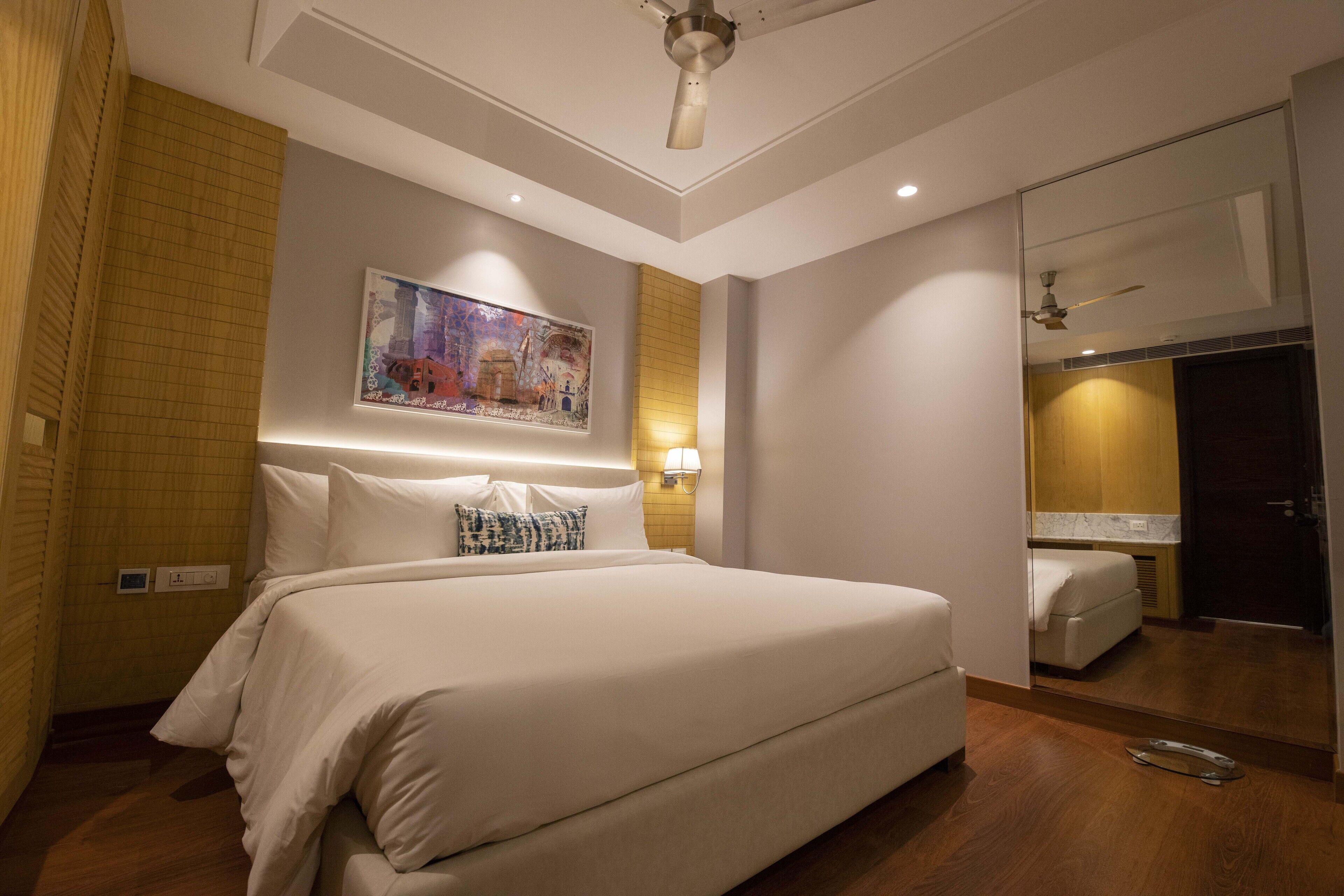 A modern hotel room with a large bed, a painting on the wall, and a mirror reflecting the room.