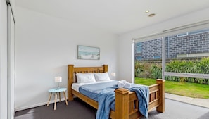 A modern bedroom with a wooden bed, a small bedside table, a nightstand, and a large window offering a view of a brick building and greenery. - A large family home situated just down the road from Port Fairy's family friendly Pea Soup Beach (PORT FAIRY)