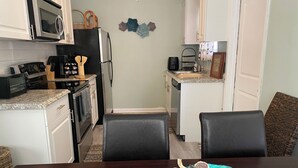 Una cocina compacta con armarios blancos, un microondas, un tostador y una cafetera. Hay una mesa de comedor con un mantel individual azul turquesa y un cuenco con caracolas.