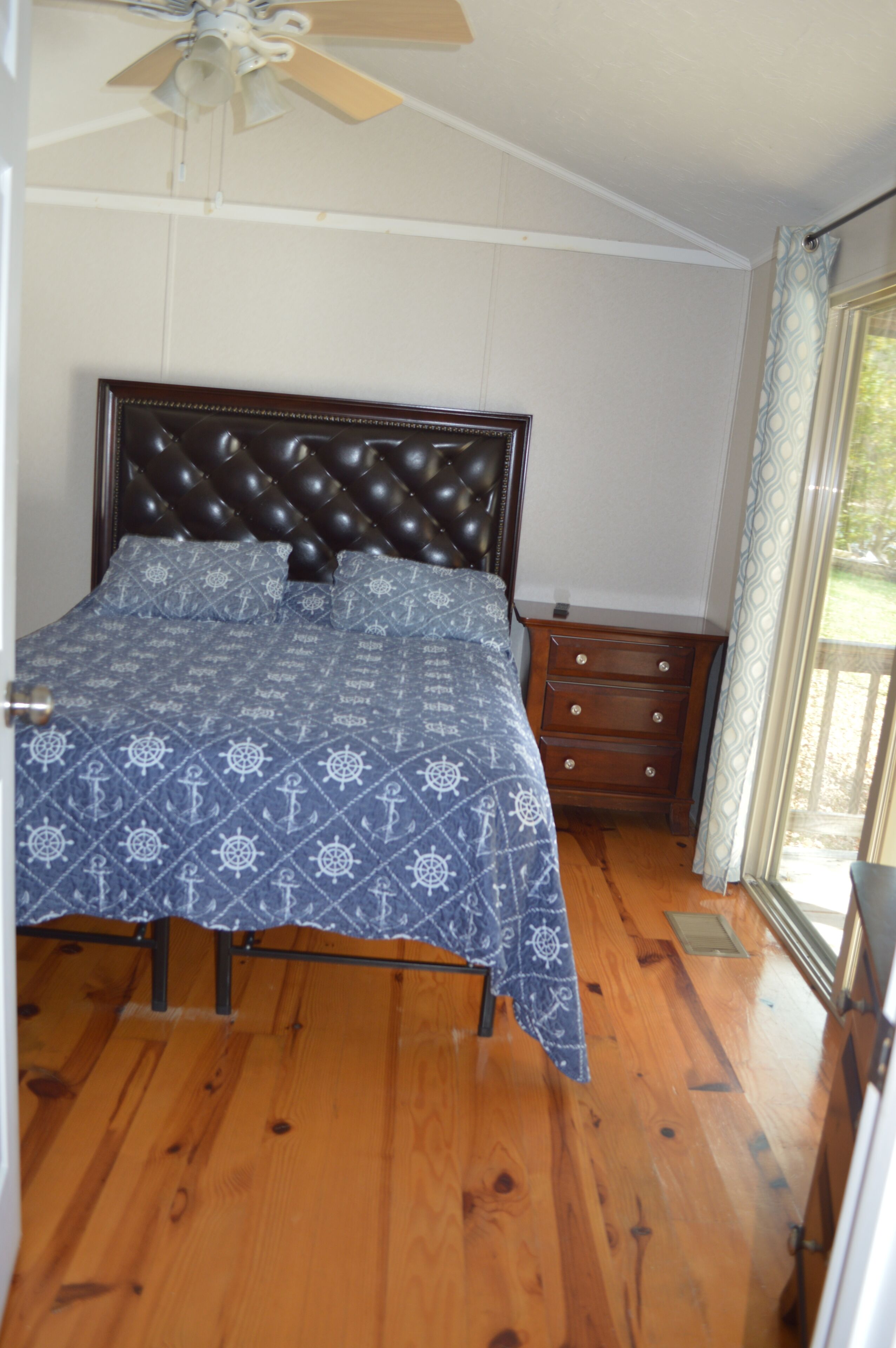 Ein Schlafzimmer mit einem Bett, einer Holzkommode, einem Deckenventilator und einem Fenster mit Vorhängen.