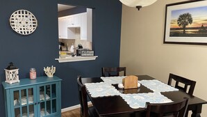 A dining area with a dark wooden table, white lace tablecloth, and a wooden block centerpiece. A blue cabinet with glass doors is to the left, and a white lantern is on top. A mirror hangs above the table, reflecting the kitchen area. - Pet Friendly Townhome Between Ocean Isle and Sunset Beach (Ocean Isle Beach)