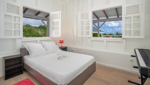 A bedroom with a bed, a nightstand, a red lamp, a window with a view of greenery, and a keyboard. - Villa Cacyli: private heated pool and Jacuzzi, view of Baie des Mulets (Le Vauclin)