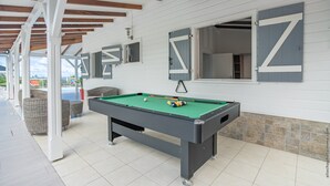 Una mesa de billar en un espacio interior, con sillas de mimbre alrededor, una vara de billar y una bola.