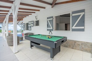 Une table de billard installée à l’intérieur, entourée de fauteuils en osier, avec une queue de billard et une bille.