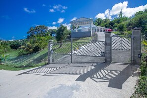 Une entrée fermée par un portail mène à un bâtiment doté d’une galerie couverte et offrant une vue sur la verdure environnante.