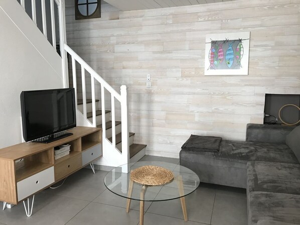 A modern living room with a wooden TV stand, a glass coffee table, a grey sofa, and a staircase with white railings.