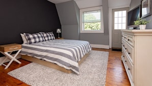 Une chambre à coucher comprenant un lit, une table de chevet, une commode et une fenêtre avec des stores.