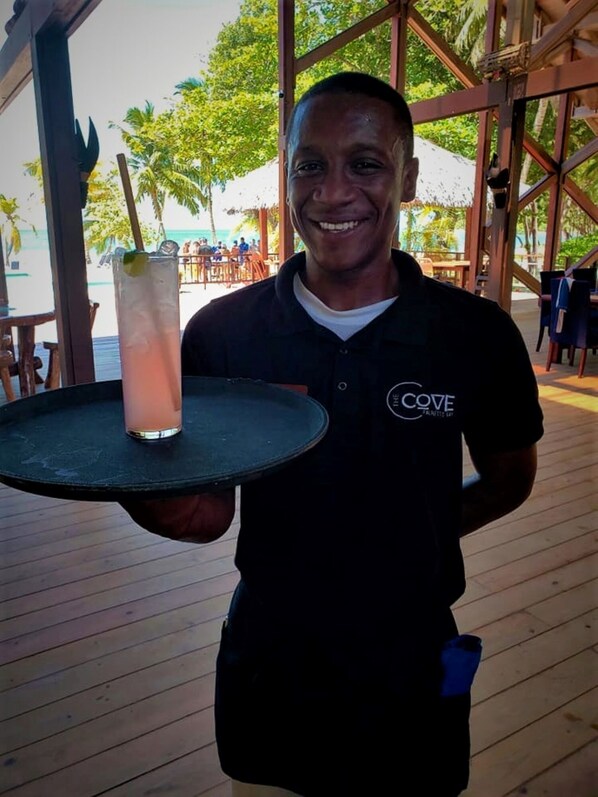A person holding a tray with a drink and a straw, wearing a black shirt with a logo, standing in an outdoor area with wooden flooring and tables, and palm trees in the background. - Cute Cottage, Private Pool, Private Beach Resort Community (Roatan)