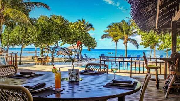 Une terrasse en bois avec une table et des chaises, donnant sur une plage bordée de palmiers et sous un ciel bleu azur.