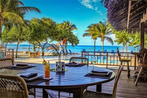 A wooden deck with a table and chairs overlooking a beach with palm trees and a clear blue sky. - Cute Cottage, Private Pool, Private Beach Resort Community (Roatan)