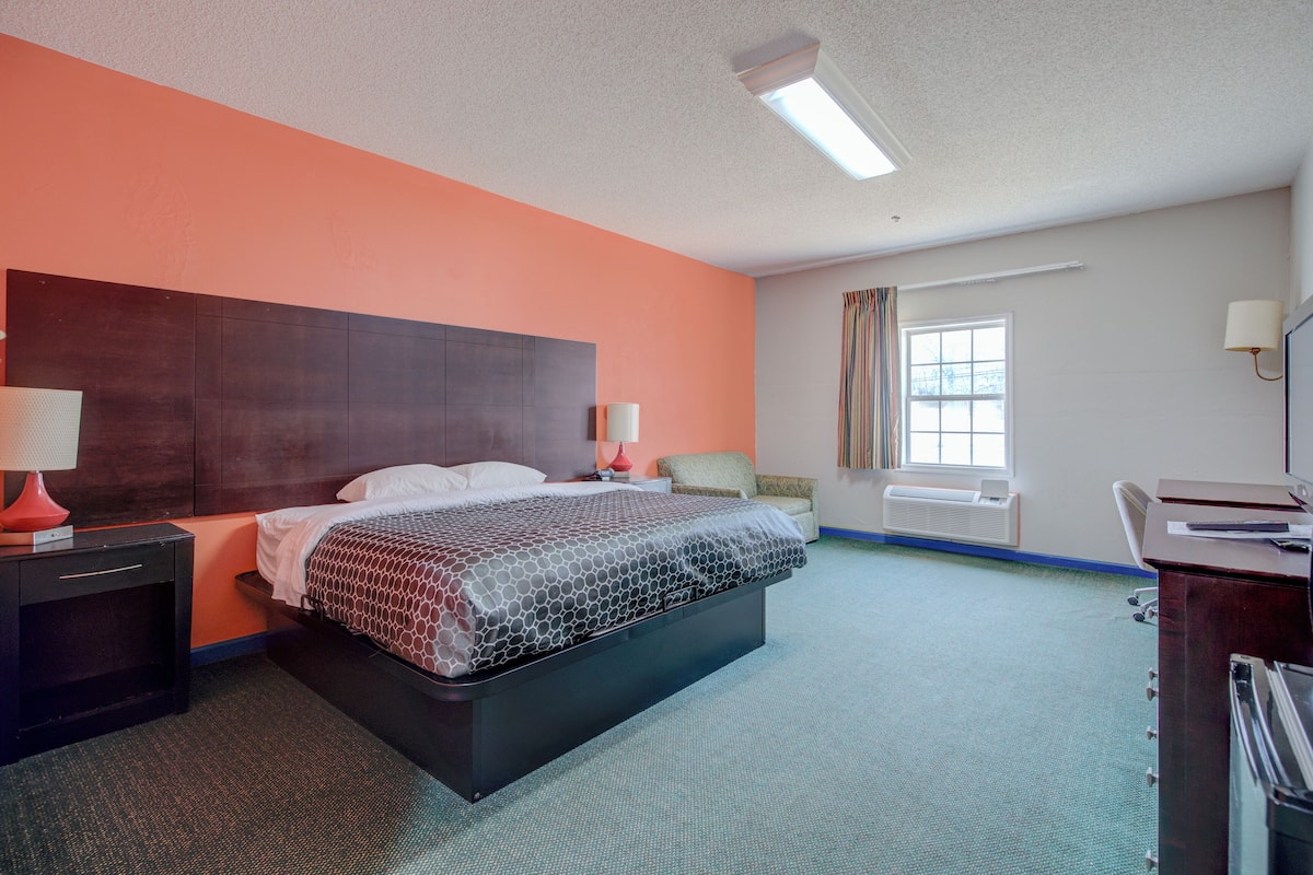 A hotel room with a bed, bedside tables, a desk, a chair, a window with curtains, and a wall-mounted lamp.