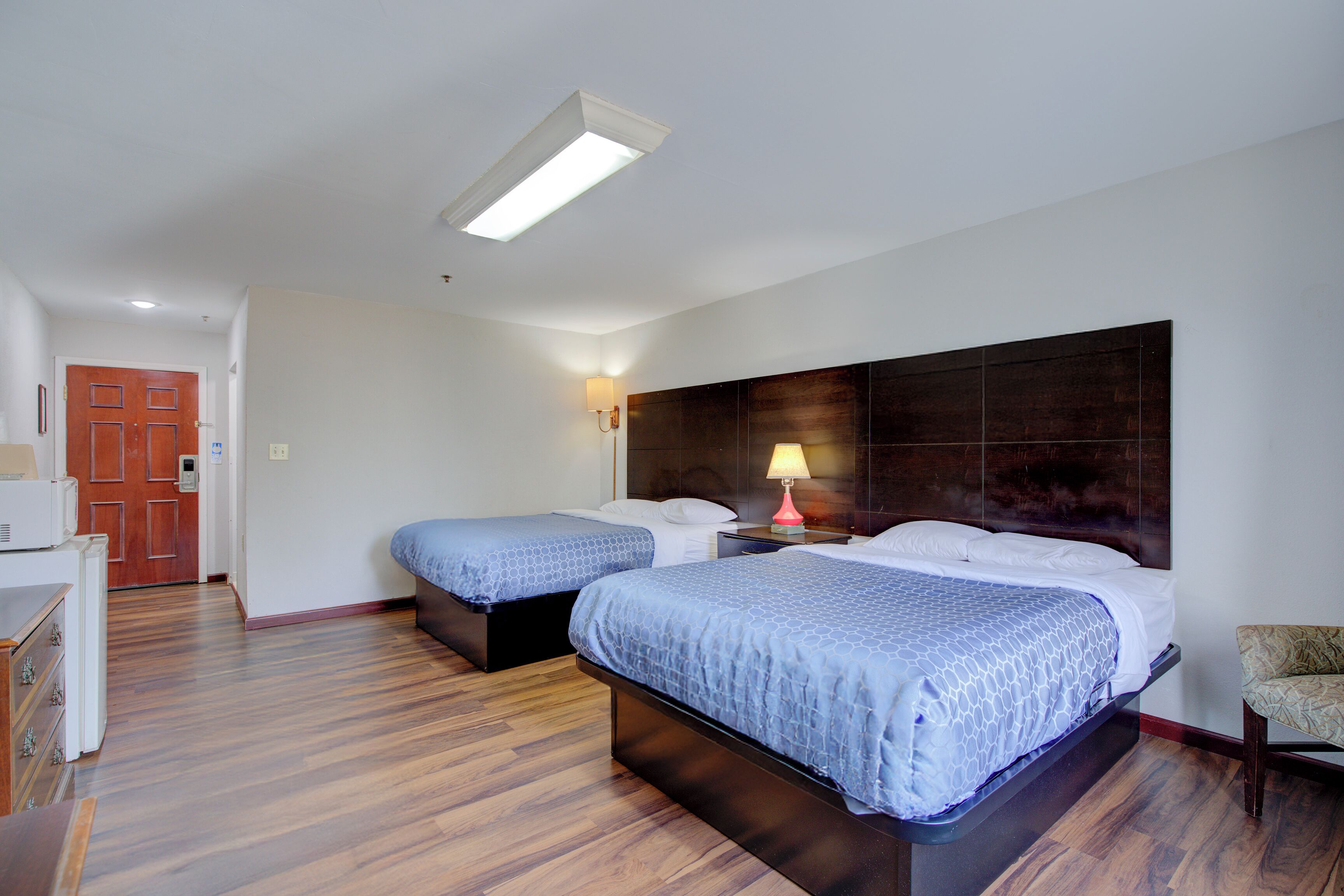 A hotel room with two beds, wooden flooring, a nightstand with a lamp, a chair, and a door.