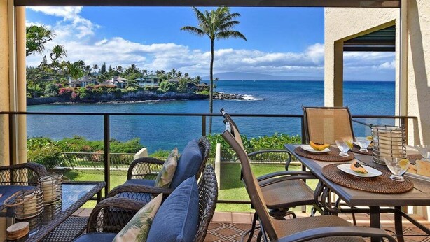 Un balcon agrémenté de meubles en osier, une table dressée avec des assiettes et des verres, et une vue sur l’océan et un paysage tropical.