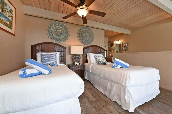 A hotel room with two beds, a ceiling fan, decorative wall art, a bedside table with a lamp, and a mirror.