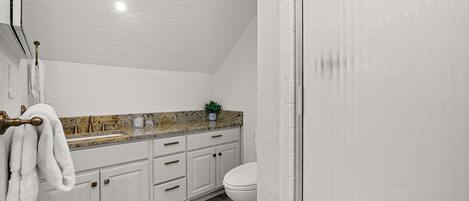 A bathroom with white cabinets, a granite countertop, a white toilet, and a shower area.