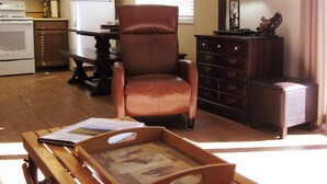 Una sala de estar con un sillón de cuero marrón, una mesa de centro de madera con una bandeja, una mesa de comedor con sillas, una zona de cocina con un refrigerador y un reloj en la pared.