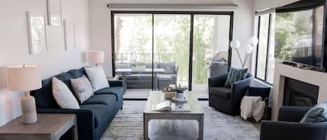A modern living room with a dark blue sofa, a coffee table, and a fireplace. There is a balcony with outdoor furniture visible through large windows.