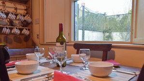 Una mesa de comedor dispuesta con fuentes, copas de vino y cubiertos, junto con una botella de vino y vistas al exterior.