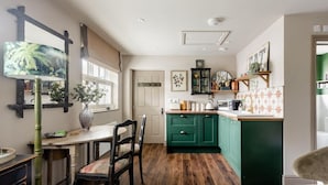 A kitchen with a wooden floor, green cabinets, a dining table with chairs, and a window with blinds. - Peaceful Studio Flat With Patio and Dogs Allowed (Cobham)