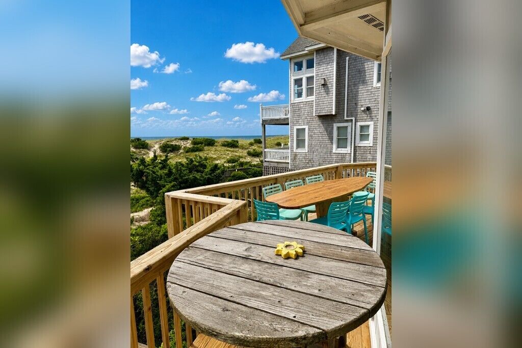 A wooden deck with a table and chairs, overlooking a beach and a building with a balcony.