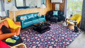 Una sala de estar con un sofá azul, un sillón amarillo y un sillón negro. El techo es de madera y hay una ventana con vista a casas.