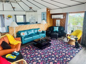 A living room with a blue sofa, a yellow armchair, and a black armchair. There is a wooden ceiling and a window with a view of houses.