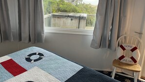 Una habitación con un ventanal, vistas a un edificio y vegetación, una cama con colcha y una silla con cojín de salvavidas.