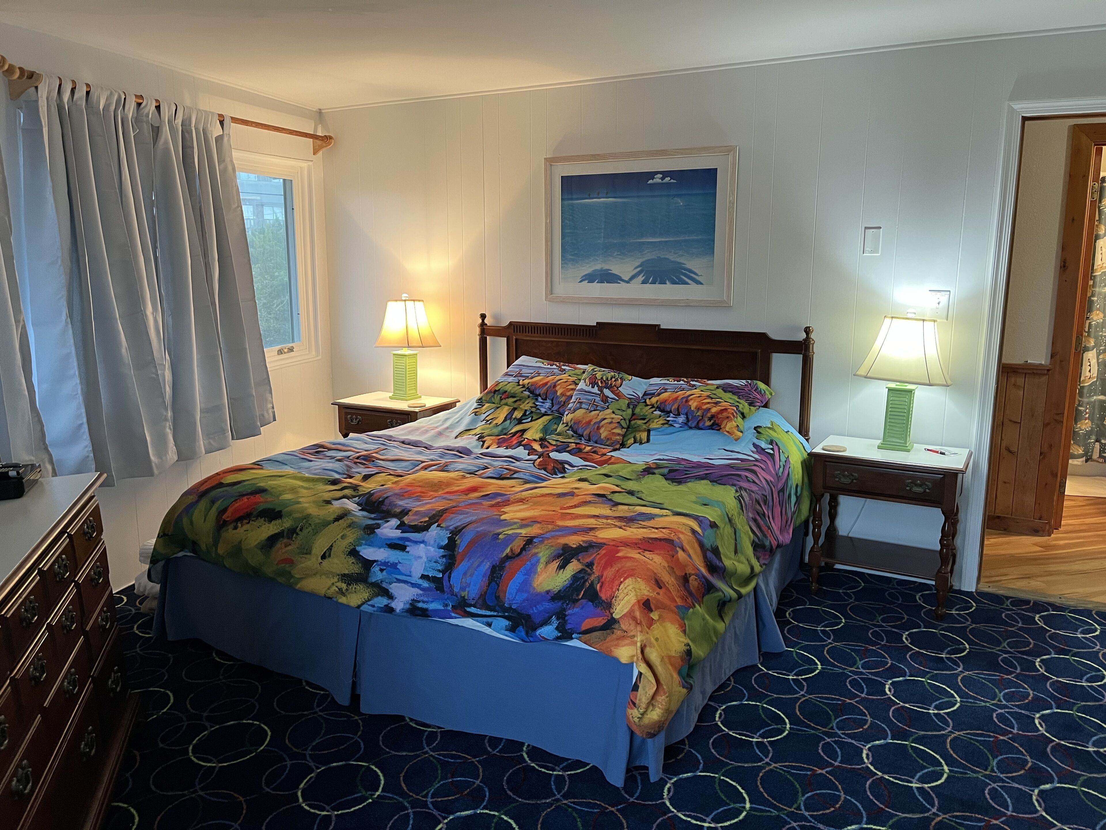 A hotel room with a large bed, two bedside tables with lamps, a wooden headboard, a painting on the wall, and a window with curtains.