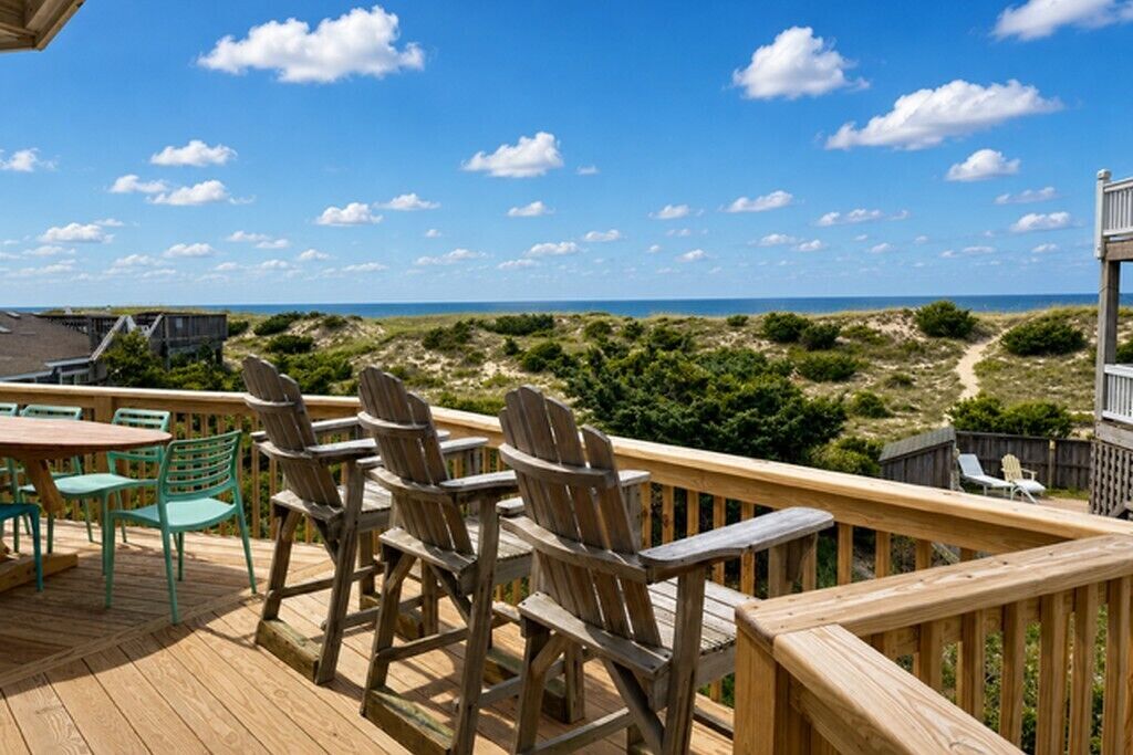 A wooden deck with outdoor seating and a view of the beach and dunes.